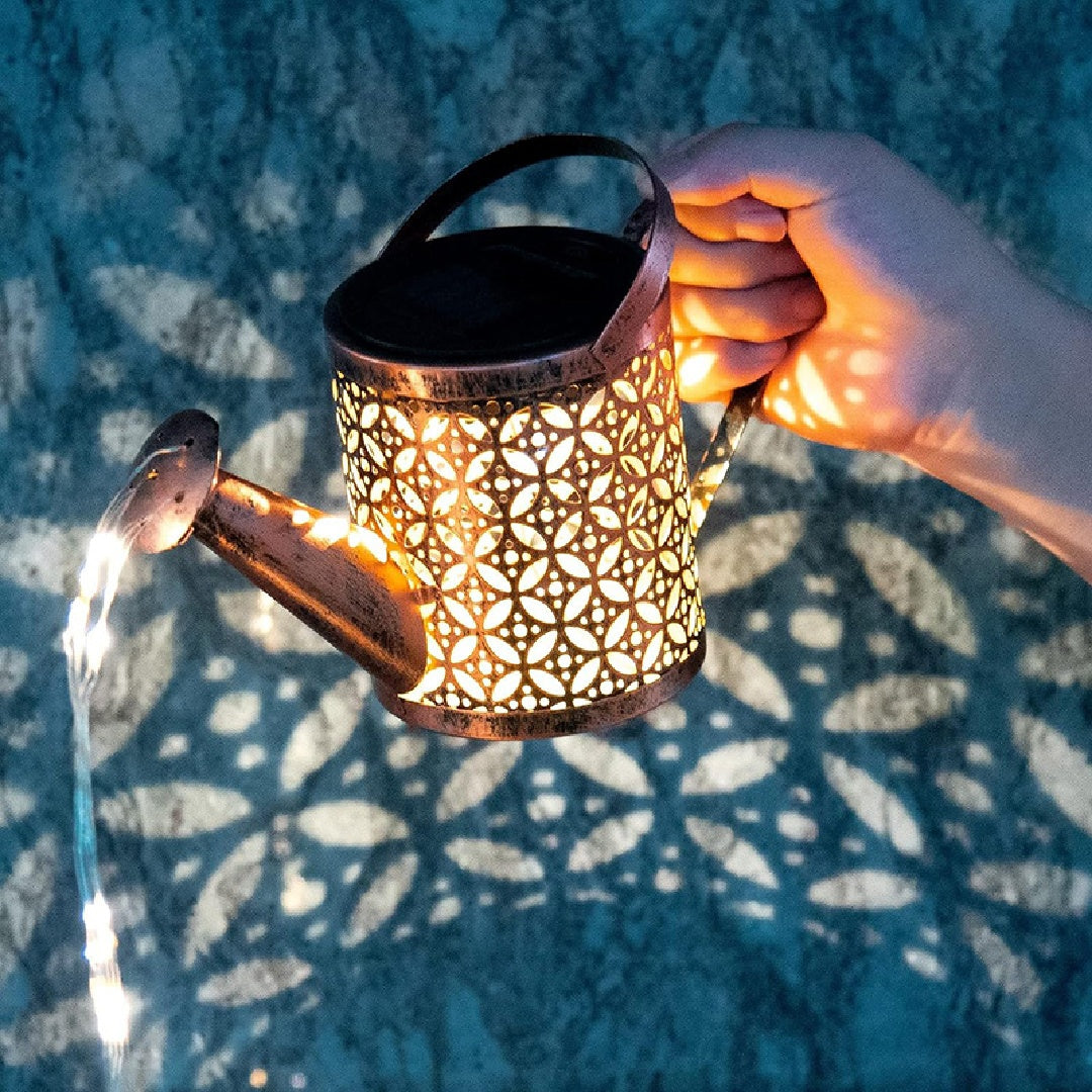 Solar-powered lantern shaped like a watering can with intricate patterns, held by a hand against a textured blue background.